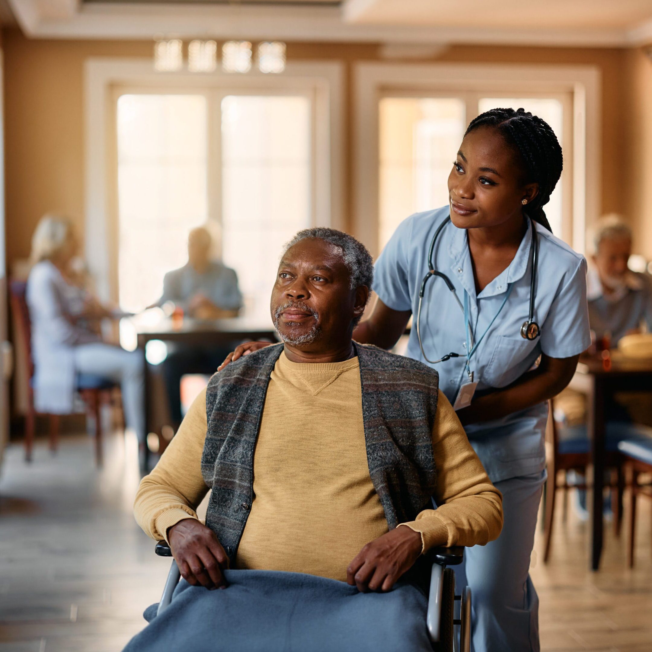 African American senior in wheelchair and young nurse looking through the window at residential care home.